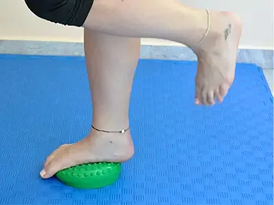 A woman stands on one leg on a green balance cushion with the other leg bent back