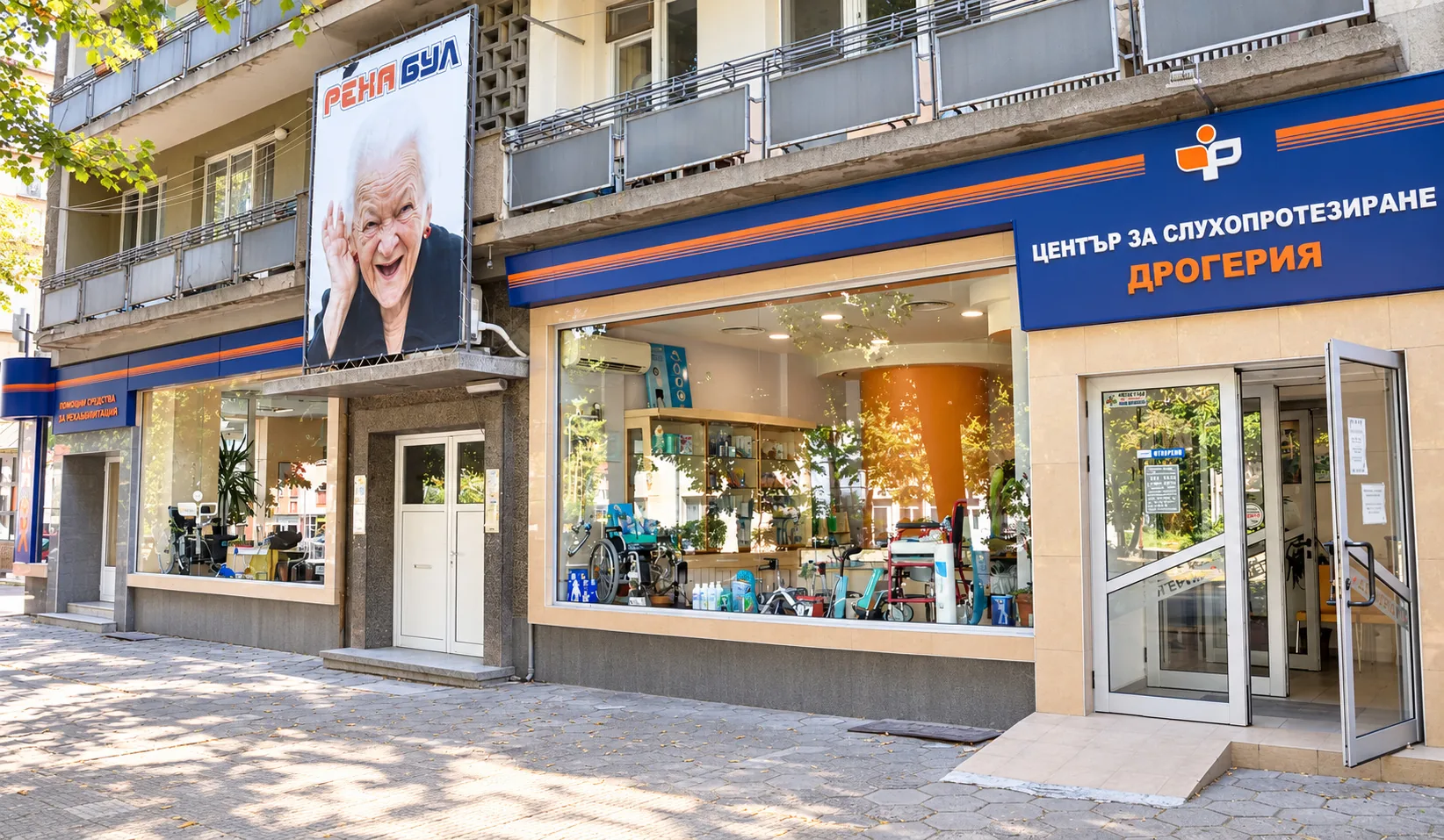 The Reha Bul shop in Gabrovo from outside: a blue sign that reads “Hearing aid centre, drugstore” in Bulgarian, a large window with assistive devices, a wide entrance and a “Reha Bul” advertising poster on the façade